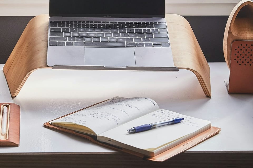 A laptop and open notebook on a wooden workspace beside a window.